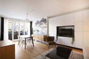 A modern living room with a dining area, a flat-screen TV mounted on a white wall, and a large window with a view of trees. - Havre de paix avec balcon (Paris)