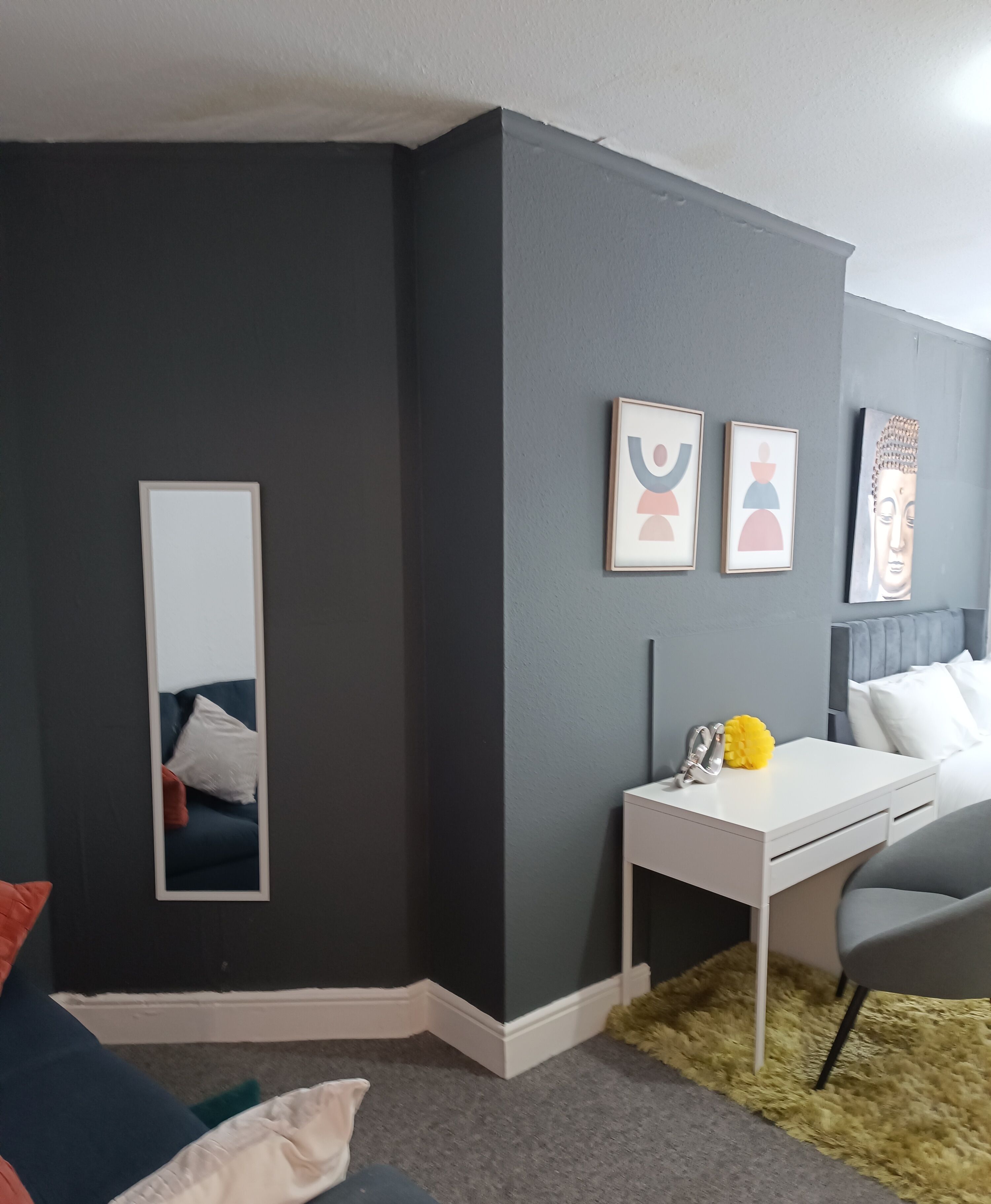 Un dormitorio moderno con paredes grises oscuras, un escritorio blanco, una silla gris y un espejo grande. Hay cuadros enmarcados en la pared y una estatua de Buda.