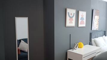 Un dormitorio moderno con paredes grises oscuras, un escritorio blanco, una silla gris y un espejo grande. Hay cuadros enmarcados en la pared y una estatua de Buda.