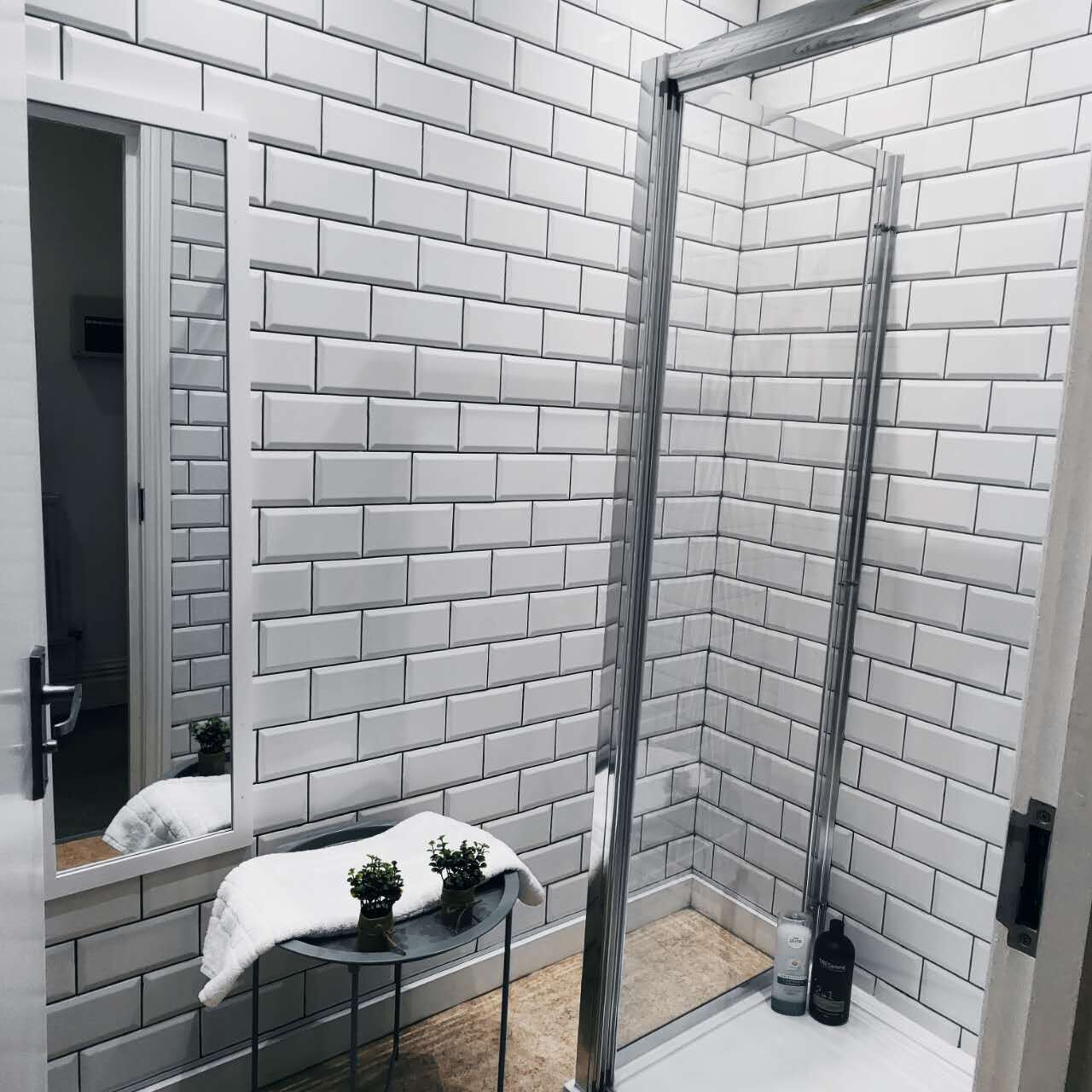 A bathroom with white brick walls, a glass shower enclosure, a small table with a towel and potted plants, and a mirror.