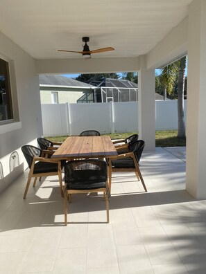 Relax on the covered patio with a wooden table and six comfortable chairs, surrounded by a white fence and a house.