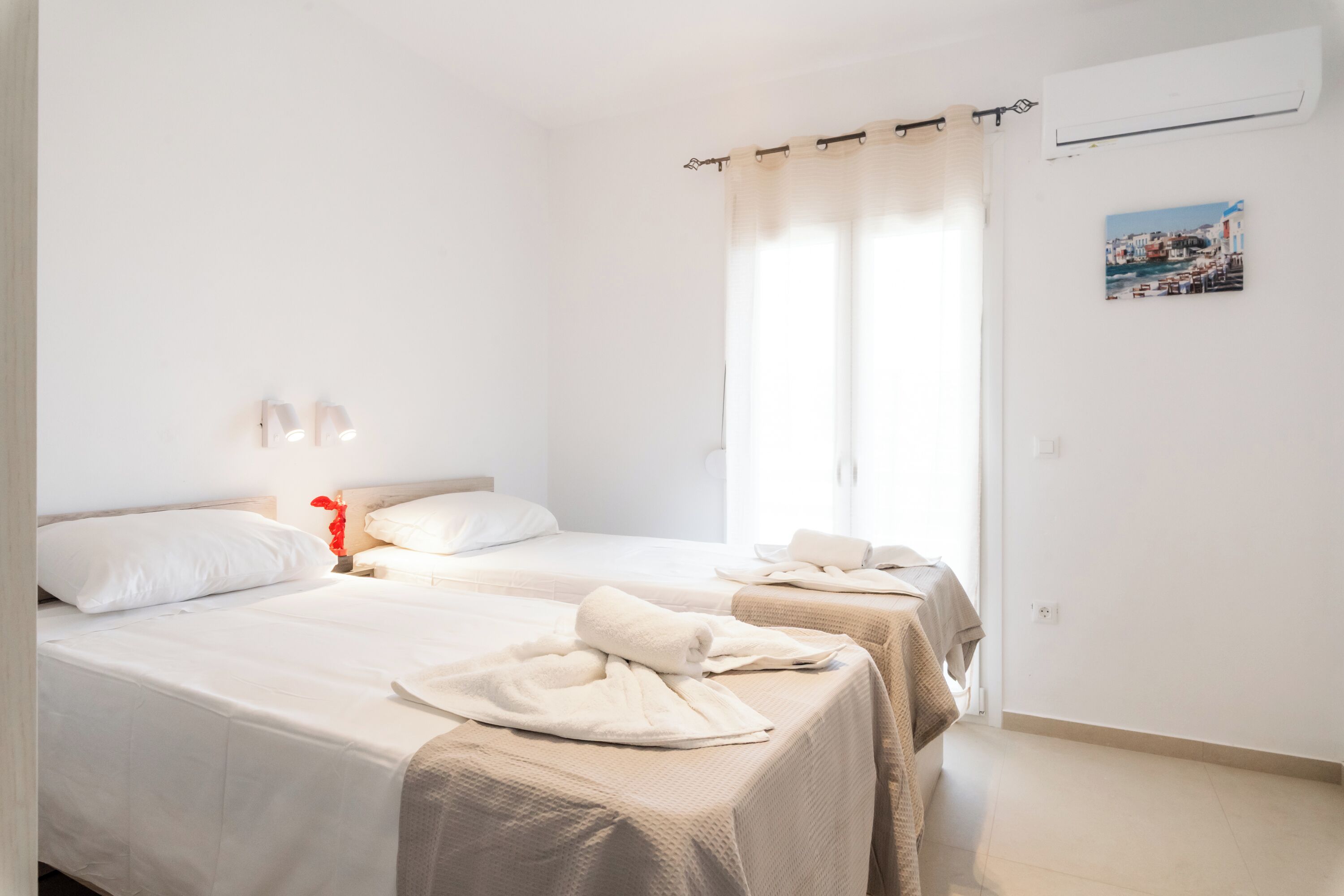 A hotel room with two beds, a window with curtains, a wall-mounted air conditioner, and a small picture on the wall.