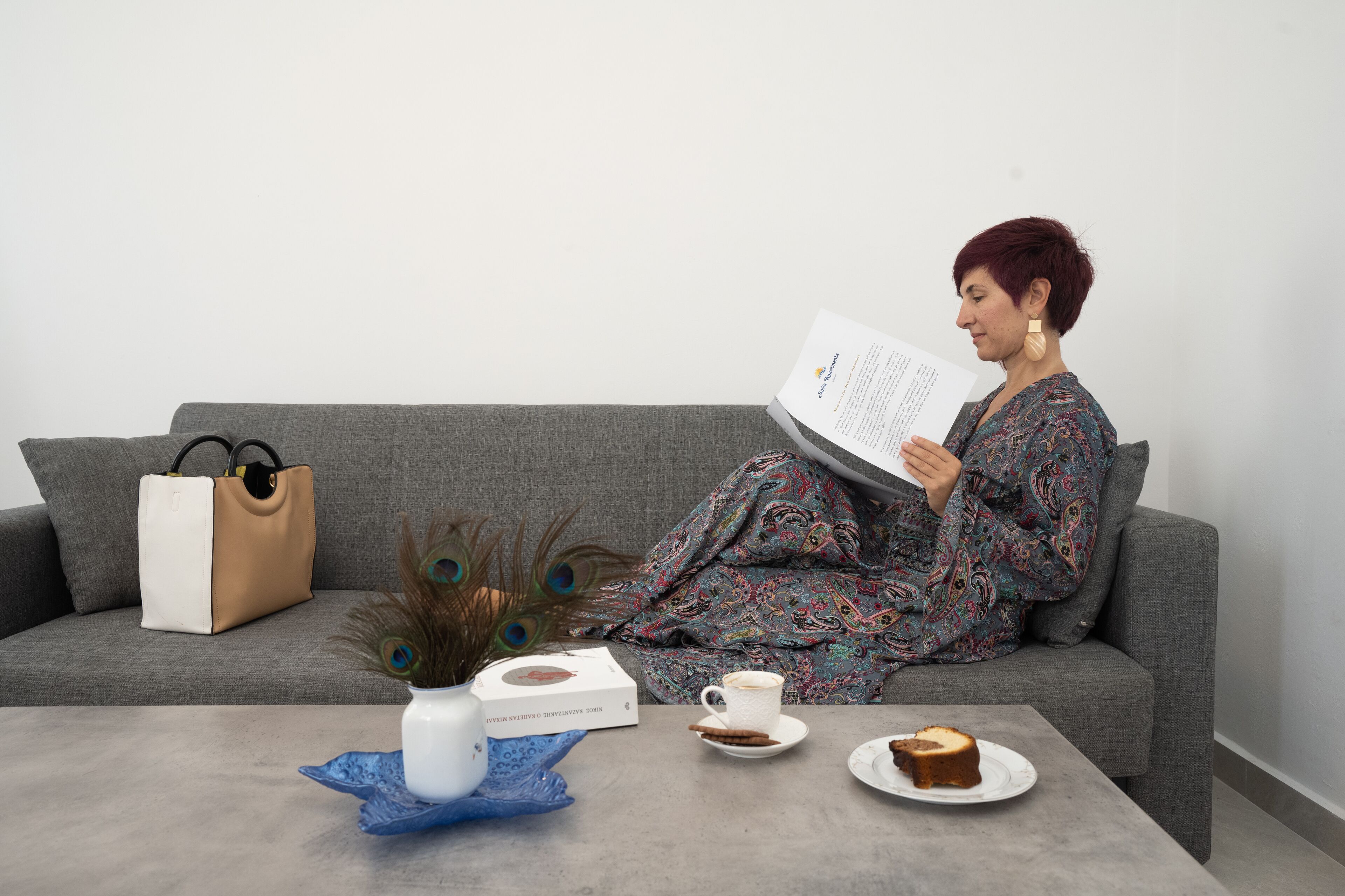 Una persona sentada en un sofá gris, leyendo un libro. Hay un jarrón blanco con base azul, una taza blanca y un plato con una porción de pastel sobre una mesa gris. Una bolsa marrón y una caja blanca están sobre el sofá.