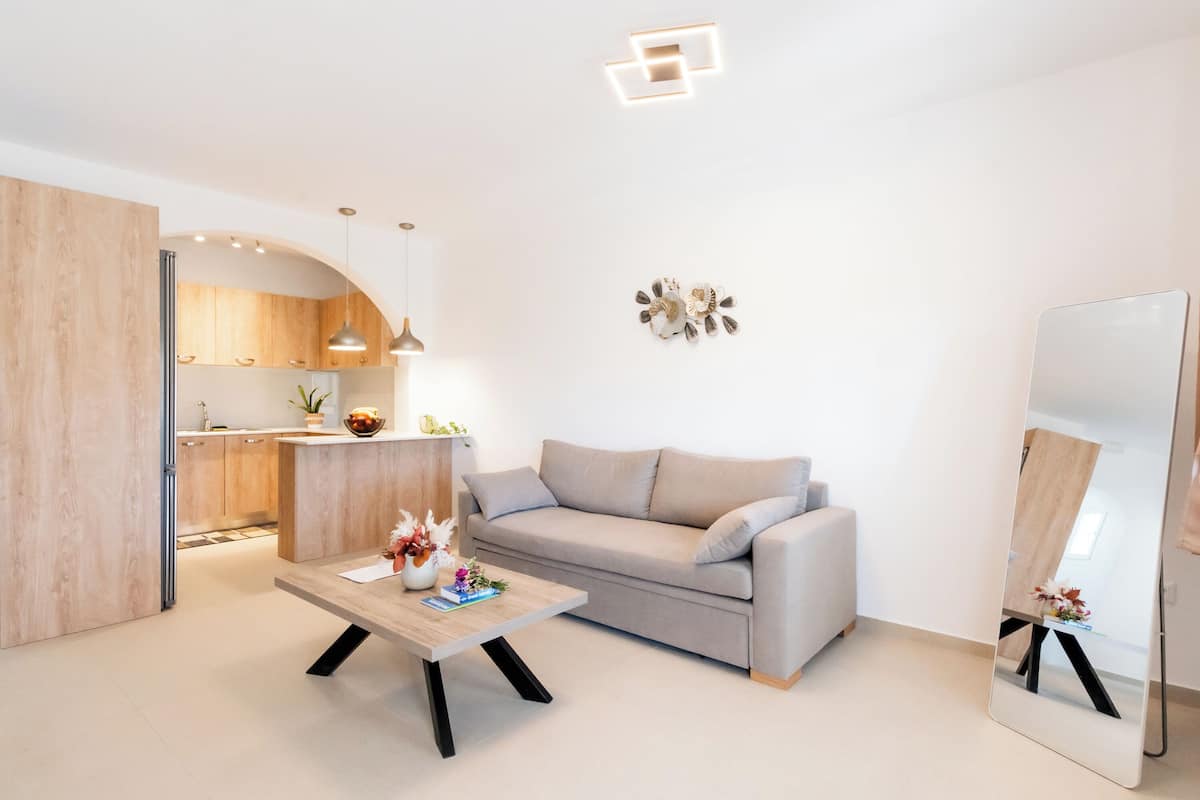 Ein modernes Wohnzimmer mit Sofa, Couchtisch, Spiegel und Küchenbereich mit Holzausbau und Pendelleuchten.