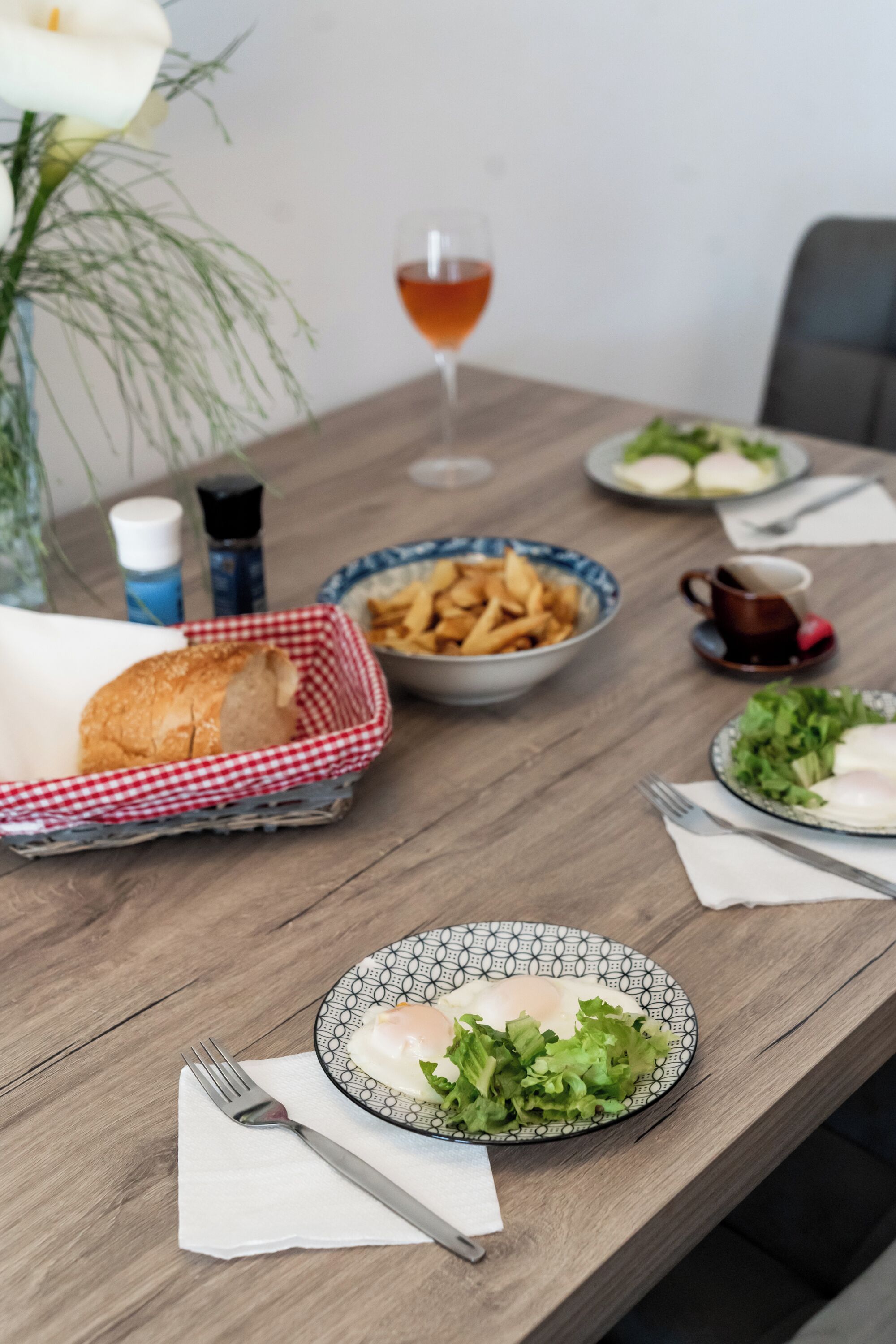 A dining table with a plate of eggs, a bowl of fries, a glass of wine, and a basket of bread.