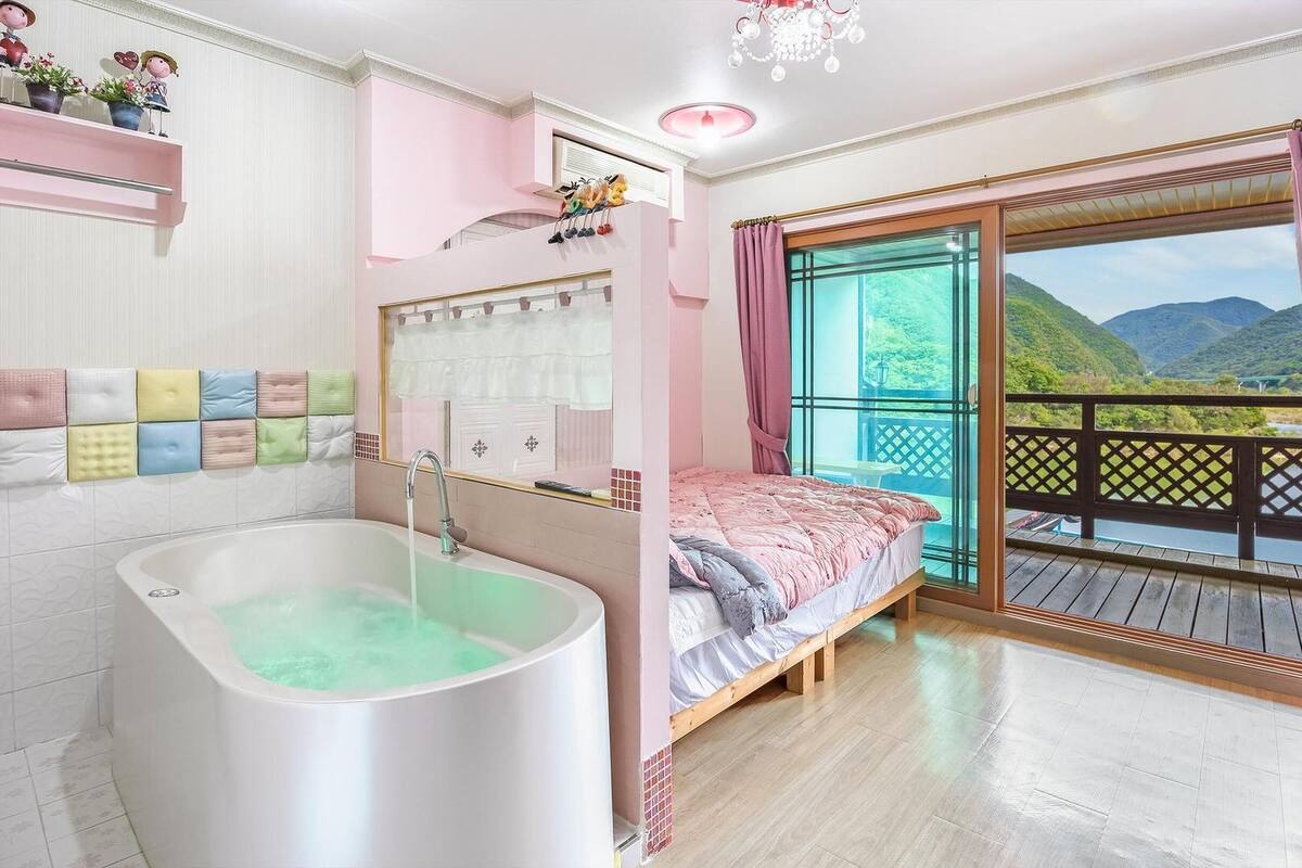 A bedroom with a large window, a bathtub, and a balcony with a view of the mountains.