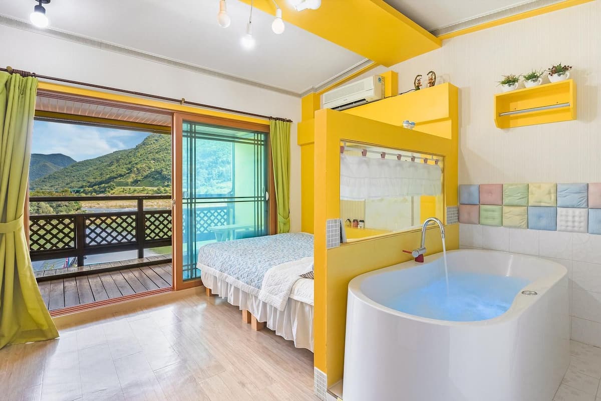 A room with a balcony, a large bathtub, and a window with a view of green hills.