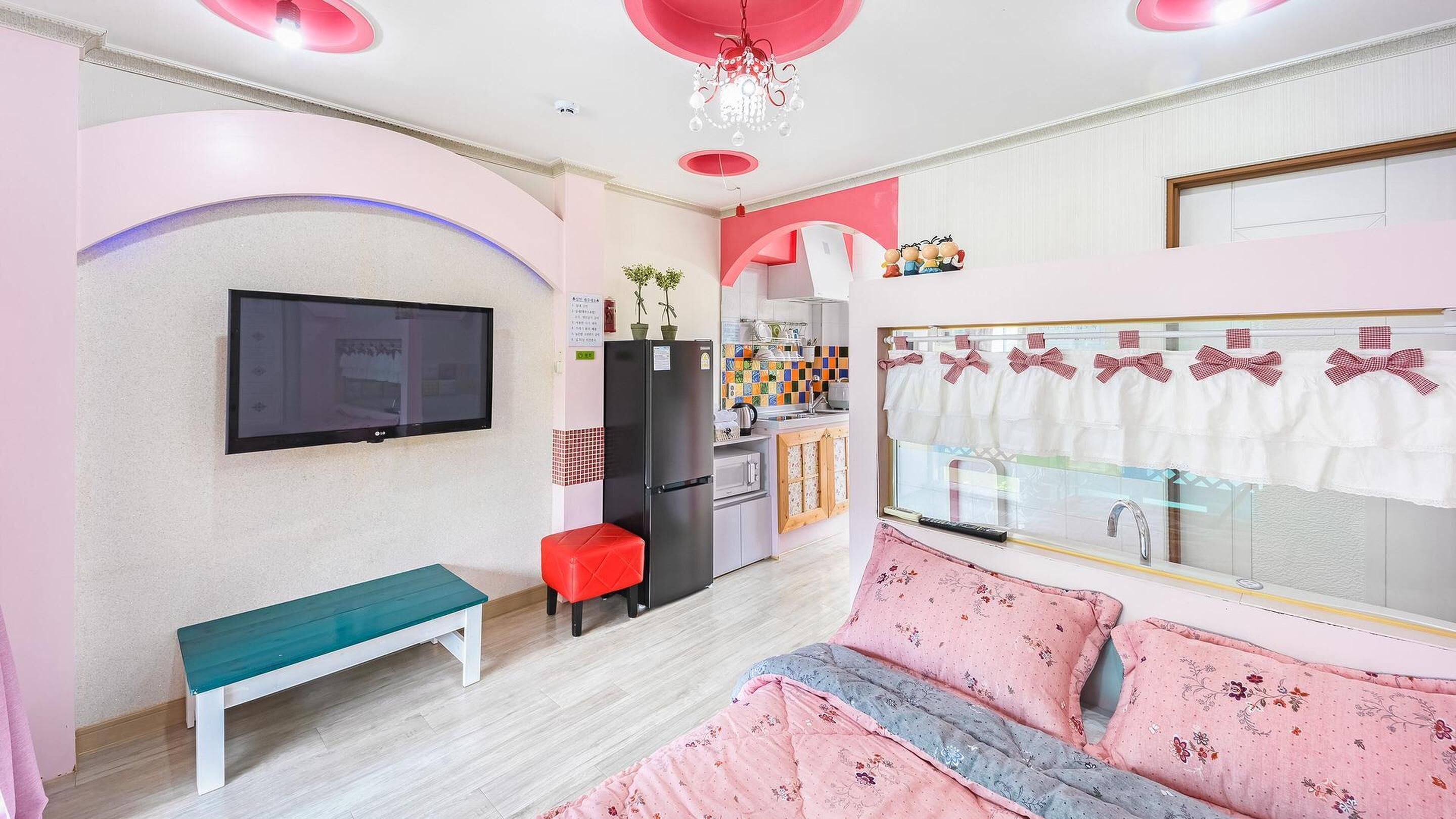 Un dormitorio moderno con televisión, una banca, una cama con ropa de cama floral y una cocina visible a través de una puerta abierta.