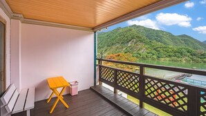 A balcony with a wooden floor, a yellow folding chair, and a trash bin. It overlooks a green landscape with a river and hills.