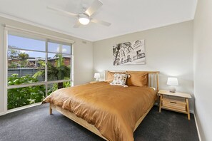 Relax in this serene bedroom with a large bed, ceiling fan, and two bedside tables with lamps, offering a view of the surrounding greenery and buildings.