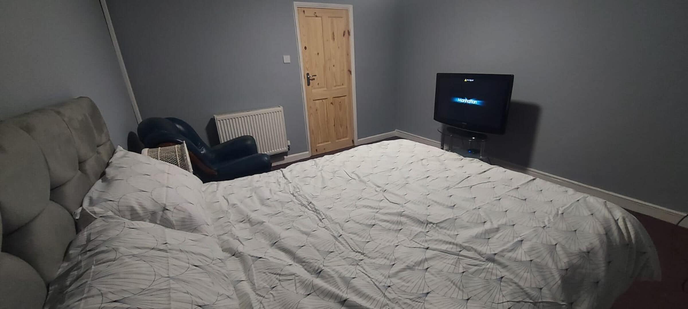 Un dormitorio con una cama grande, una silla, un radiador, una puerta de madera y un televisor.
