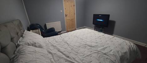 Un dormitorio con una cama grande, una silla, un radiador, una puerta de madera y un televisor.