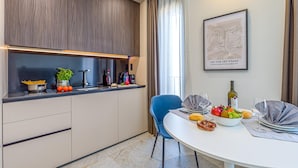 A modern kitchen with a built-in oven, a dining table set with a bowl of fruit, wine glasses, and a bottle of wine. - Icon H 405 Suite Business (Lugano)