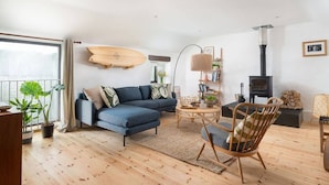 Una sala de estar moderna con un sofá azul, piso de madera, una tabla de surf en la pared, chimenea y un ventanal grande.