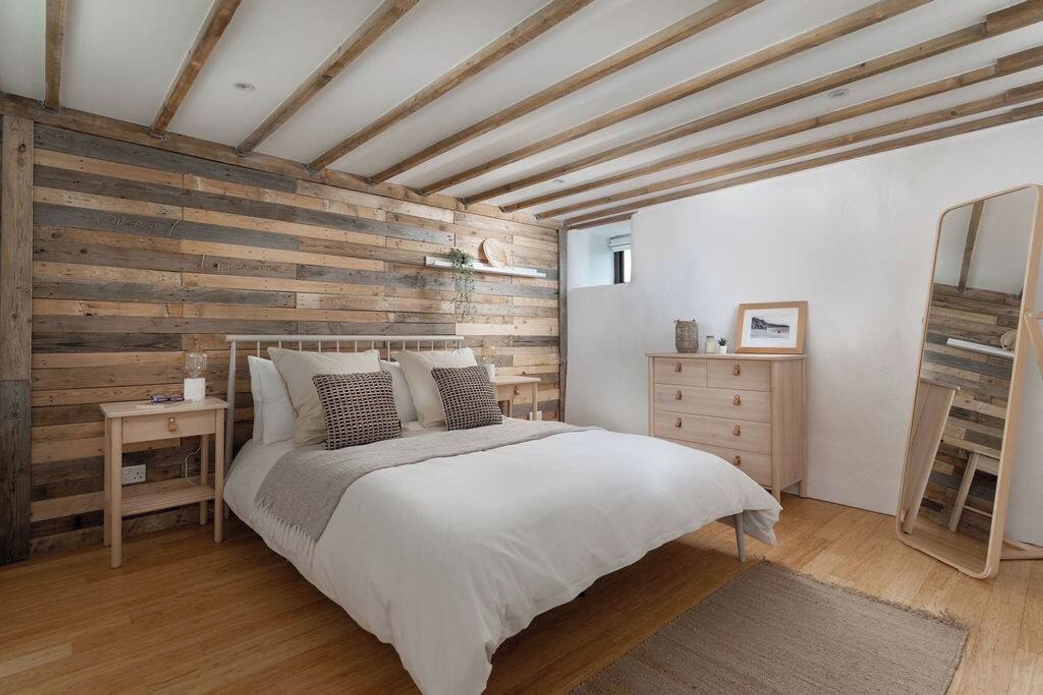 A bedroom with a wooden slat wall, a bed with white bedding, a bedside table, a dresser, and a mirror.