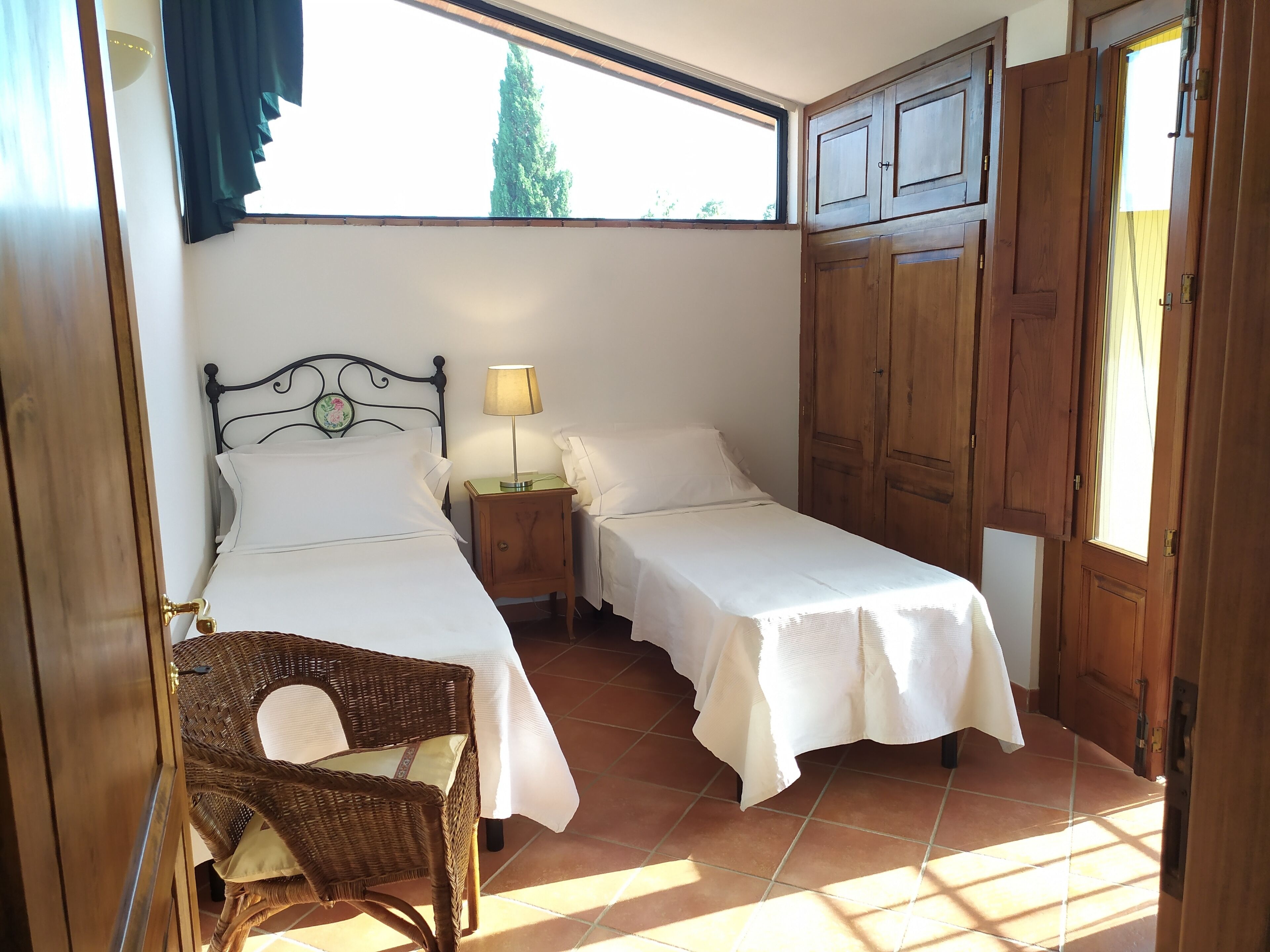 Un dormitorio con dos camas individuales, una mesita de noche con lámpara, una silla de mimbre y un armario.