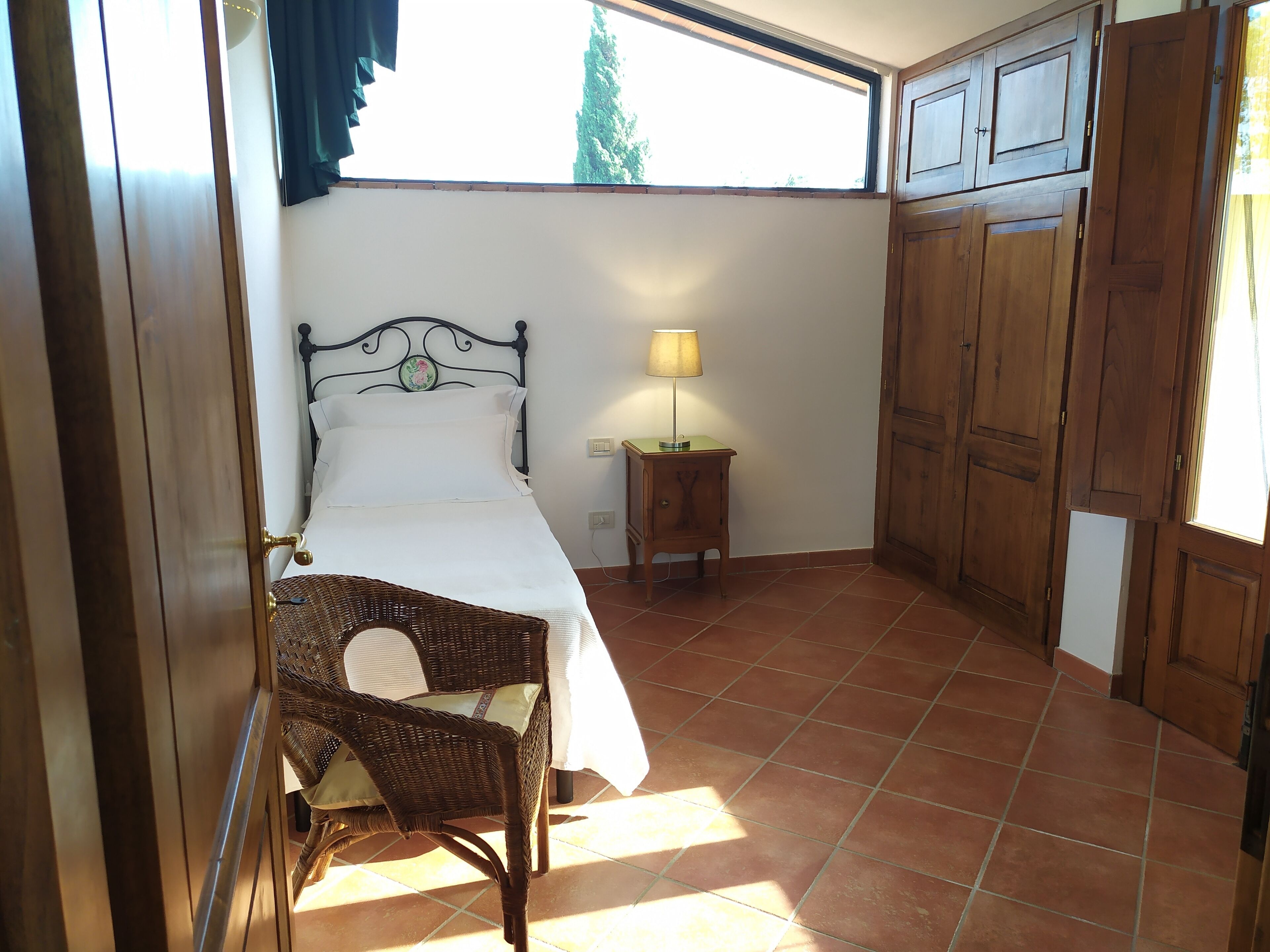 A bedroom with a bed, a nightstand, a lamp, a wicker chair, and a wardrobe.