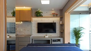 Un dormitorio moderno con una cama, un televisor montado en la pared, una kitchenette pequeña y un ventanal grande.