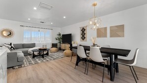 A modern living room with a gray sofa, a dining table with chairs, a TV, and decorative wall art. - Vista del Valle 🌵 Pickleball + Pitch&Putt + Hot Tub + Pool (Joshua Tree)