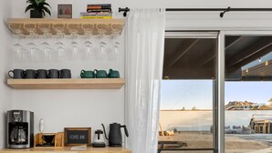 A room with a coffee station, shelves with mugs, a coffee maker, and a view through a glass door. - Vista del Valle 🌵 Pickleball + Pitch&Putt + Hot Tub + Pool (Joshua Tree)