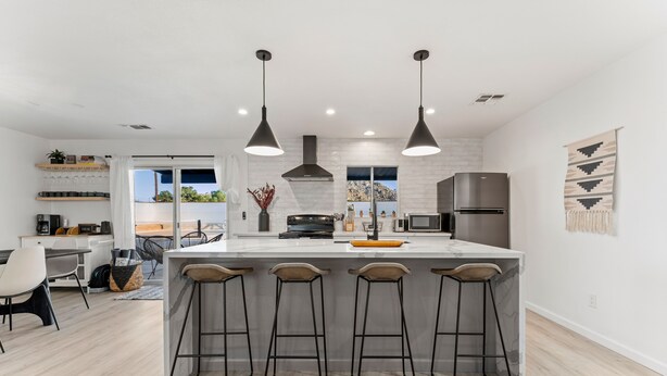 A modern kitchen with a large island, bar stools, pendant lights, and a view of the outdoors. - Vista del Valle 🌵 Pickleball + Pitch&Putt + Hot Tub + Pool (Joshua Tree)