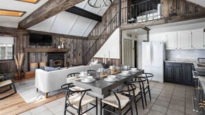 Ein großzügiger Loft-Wohnbereich mit hoher Decke, freiliegenden Holzbalken, Kamin, Esstisch für sechs Personen und einer Küche mit dunklen Schränken.