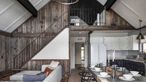 Ein geräumiger Loft mit hoher Decke, freiliegenden Holzbalken, einer Treppe, einem Essbereich mit langem Tisch und Stühlen sowie einer Küche mit weißen Schränken.