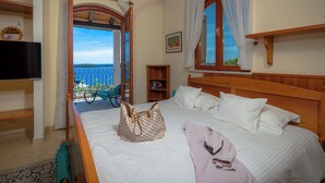A bedroom with a bed, a wooden headboard, a window with curtains, a view of the sea, a television on the wall, and a small wooden shelf.