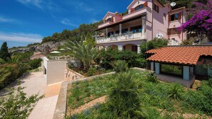 A large, two-story house with a balcony, surrounded by lush greenery and a clear blue sky.