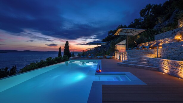 A modern infinity pool with a wooden deck, overlooking a body of water and a sunset.