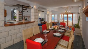 A dining area with a long table set for six, surrounded by wicker chairs with red cushions.