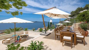 A terrace with wooden furniture, white umbrellas, and a view of the sea.