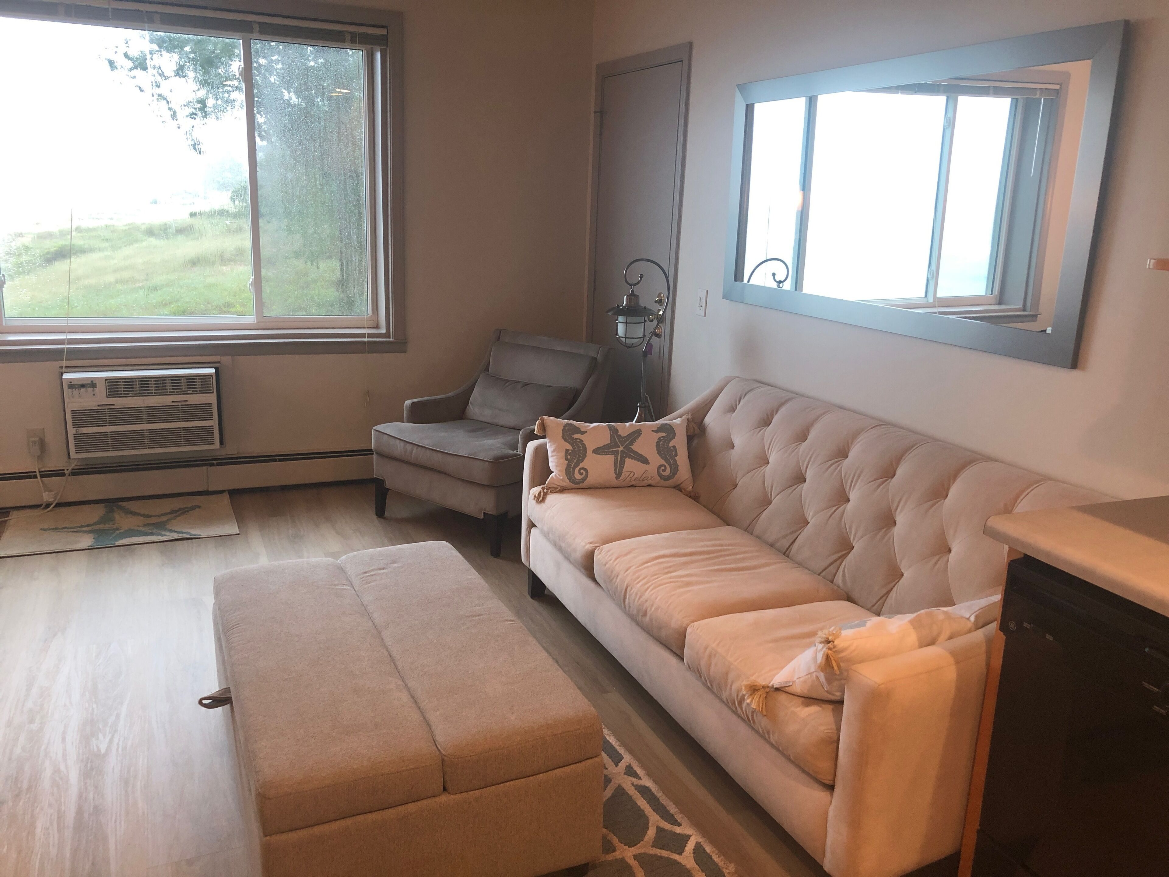 A living room with a beige sofa, a matching ottoman, a grey armchair, and a window with a view of greenery.