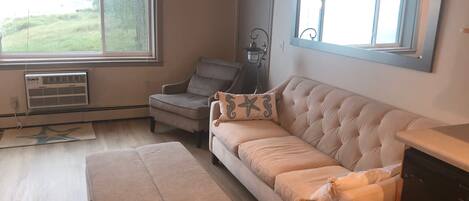 A living room with a beige sofa, a matching ottoman, a grey armchair, and a window with a view of greenery.