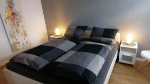 A bedroom with a bed, two bedside tables with lamps, a white chair, and a wall with a colorful abstract painting. - Funke Vacation Rental (Reichshof)