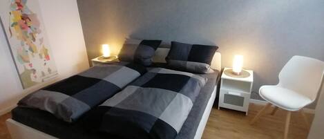 Een slaapkamer met een bed, twee nachtkastjes met lampen, een witte stoel en een muur met een kleurrijk abstract schilderij.