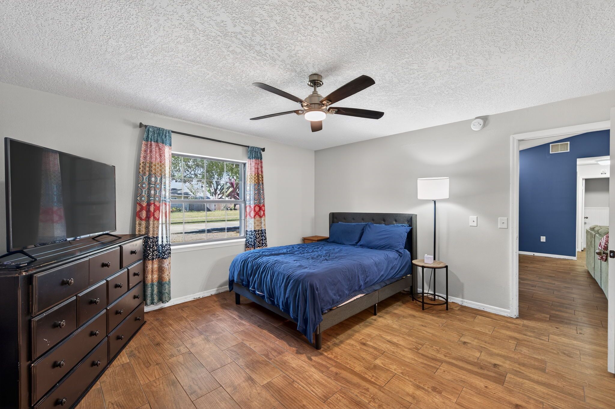 A bedroom with a bed, dresser, ceiling fan, and a window with curtains.