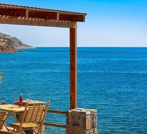 A wooden pergola overlooking a clear blue sea with a stone wall and a table with chairs and a bottle of red wine.