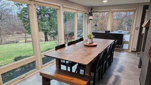 A spacious dining area with a large wooden table, black chairs, and a bench. It features large windows offering a view of a grassy yard and trees.