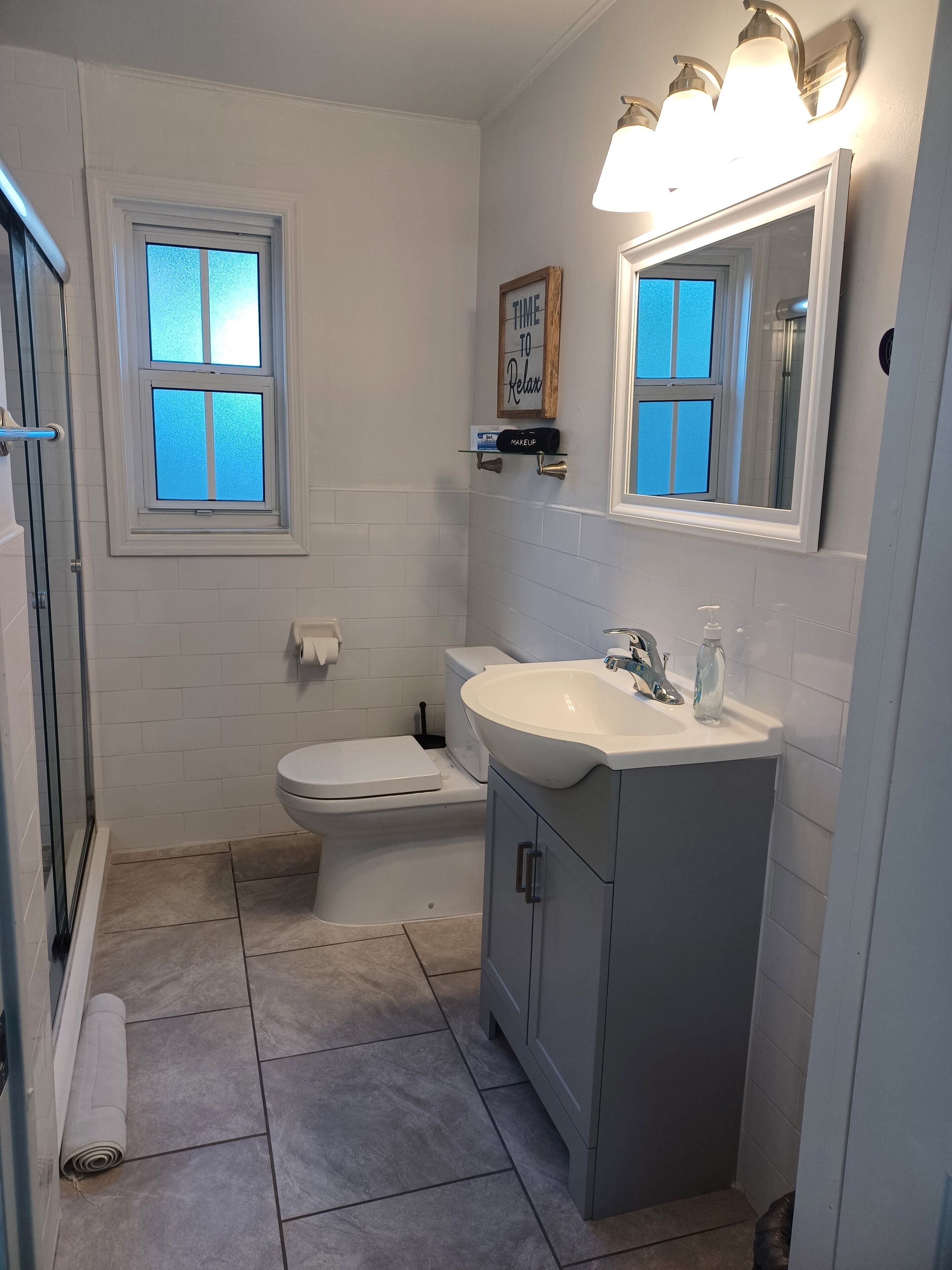 A bathroom with a toilet, sink, and shower. There is a window with frosted glass and a sign above the mirror.
