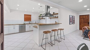 Una cocina moderna con armarios blancos, una isla central con taburetes, una campana extractora de acero inoxidable y luces empotradas en el techo.
