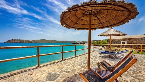 Een terras van natuursteen met lounge stoelen en een rieten parasol, met uitzicht op een turquoise zee en heuvels in de verte.