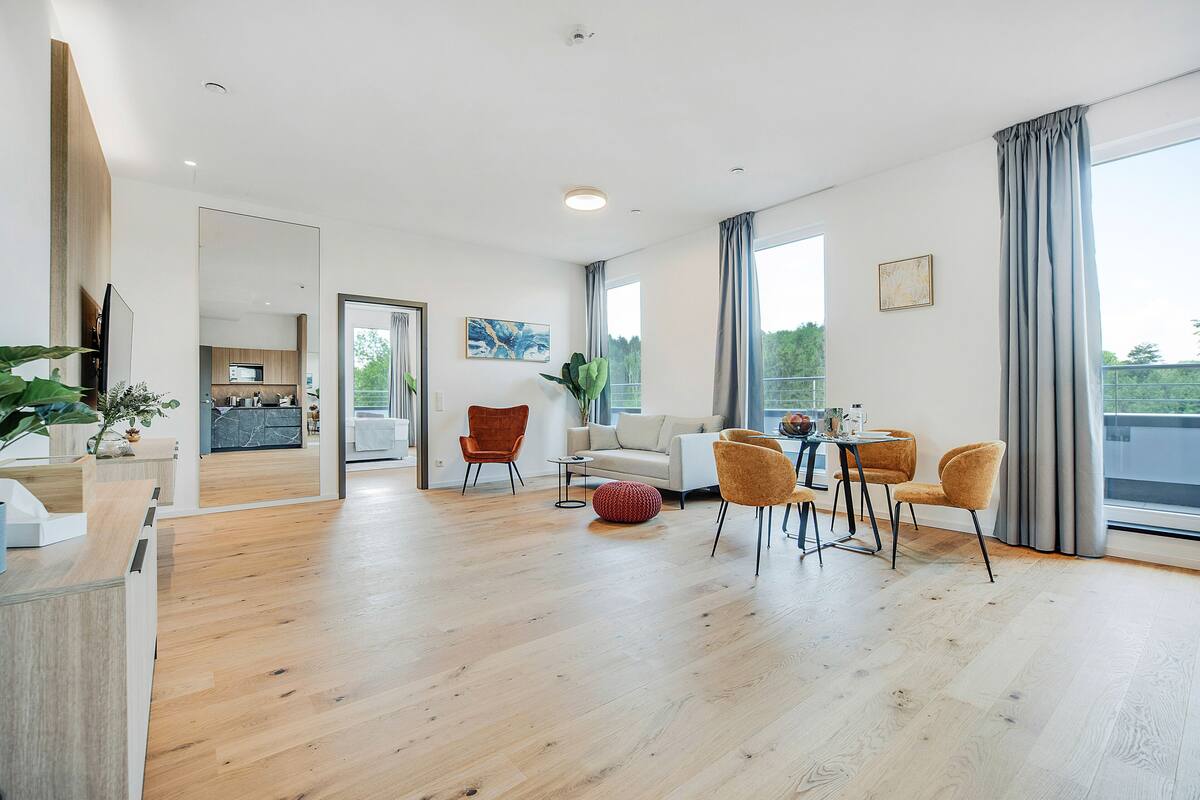 A spacious living area with a wooden floor, a sofa, two armchairs, a dining table with chairs, and large windows with curtains.