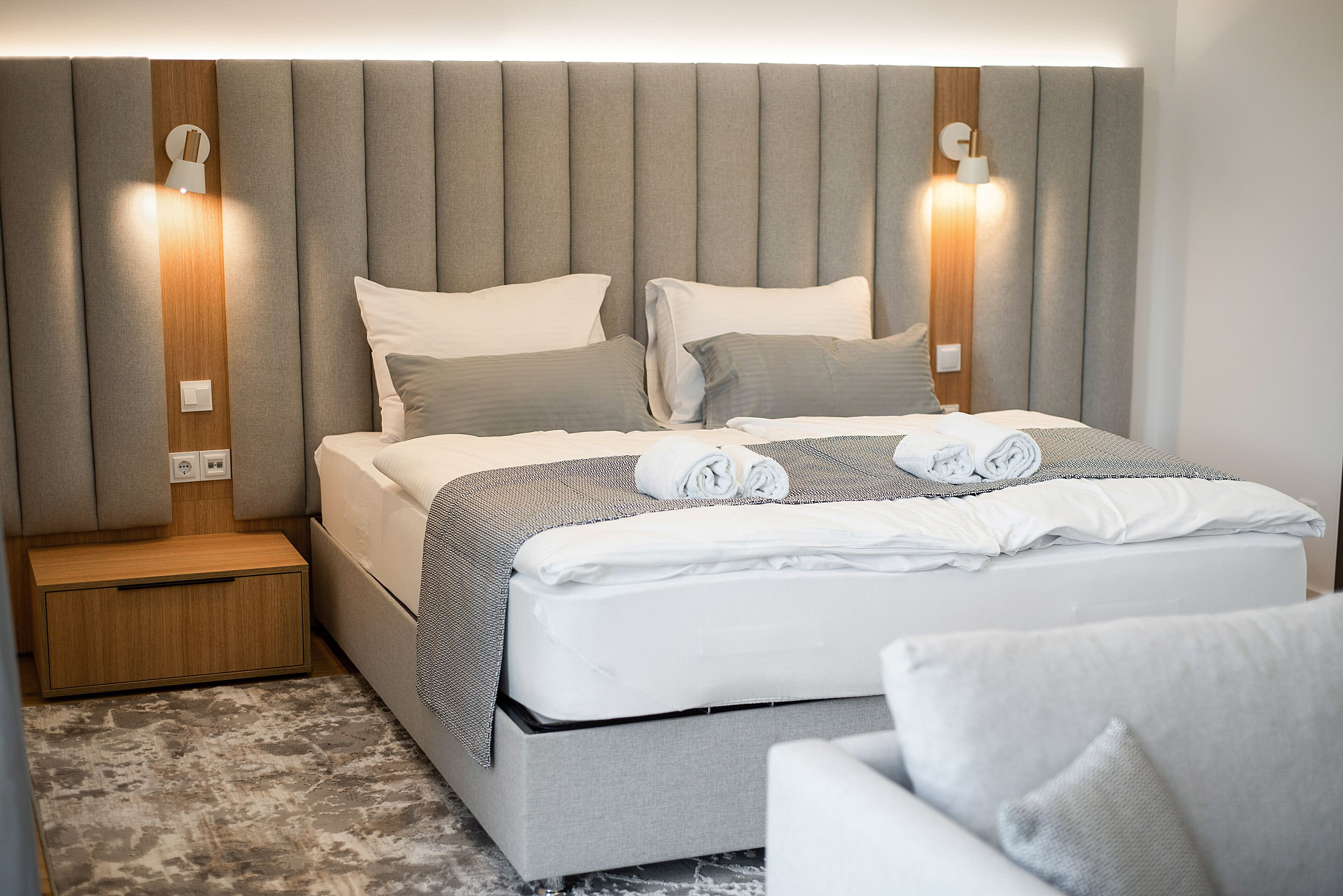 A neatly made bed with a grey headboard, two bedside lamps, and a small wooden nightstand.