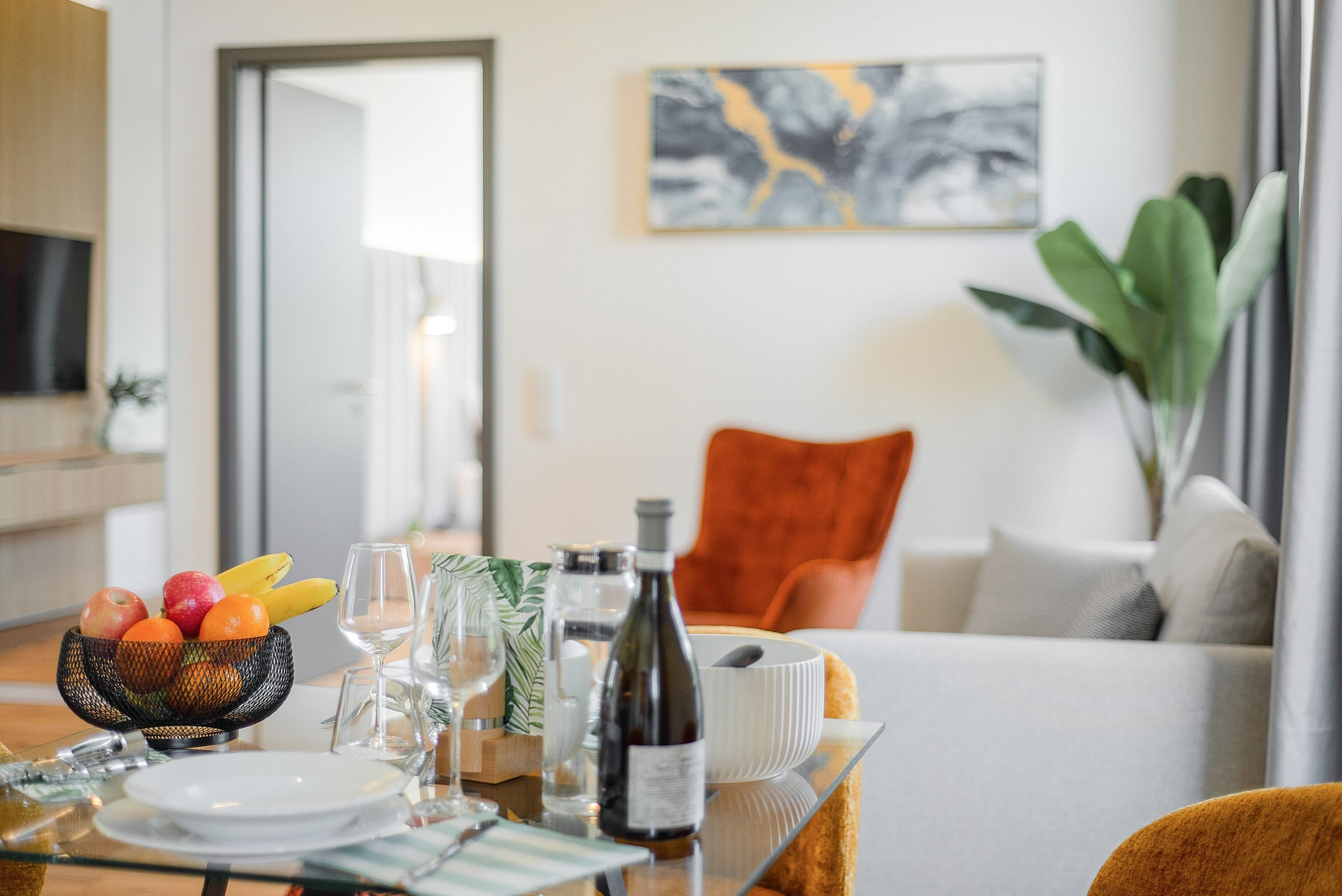 A dining table set with fruits, wine, and glasses, a sofa, and a plant in a modern living room.