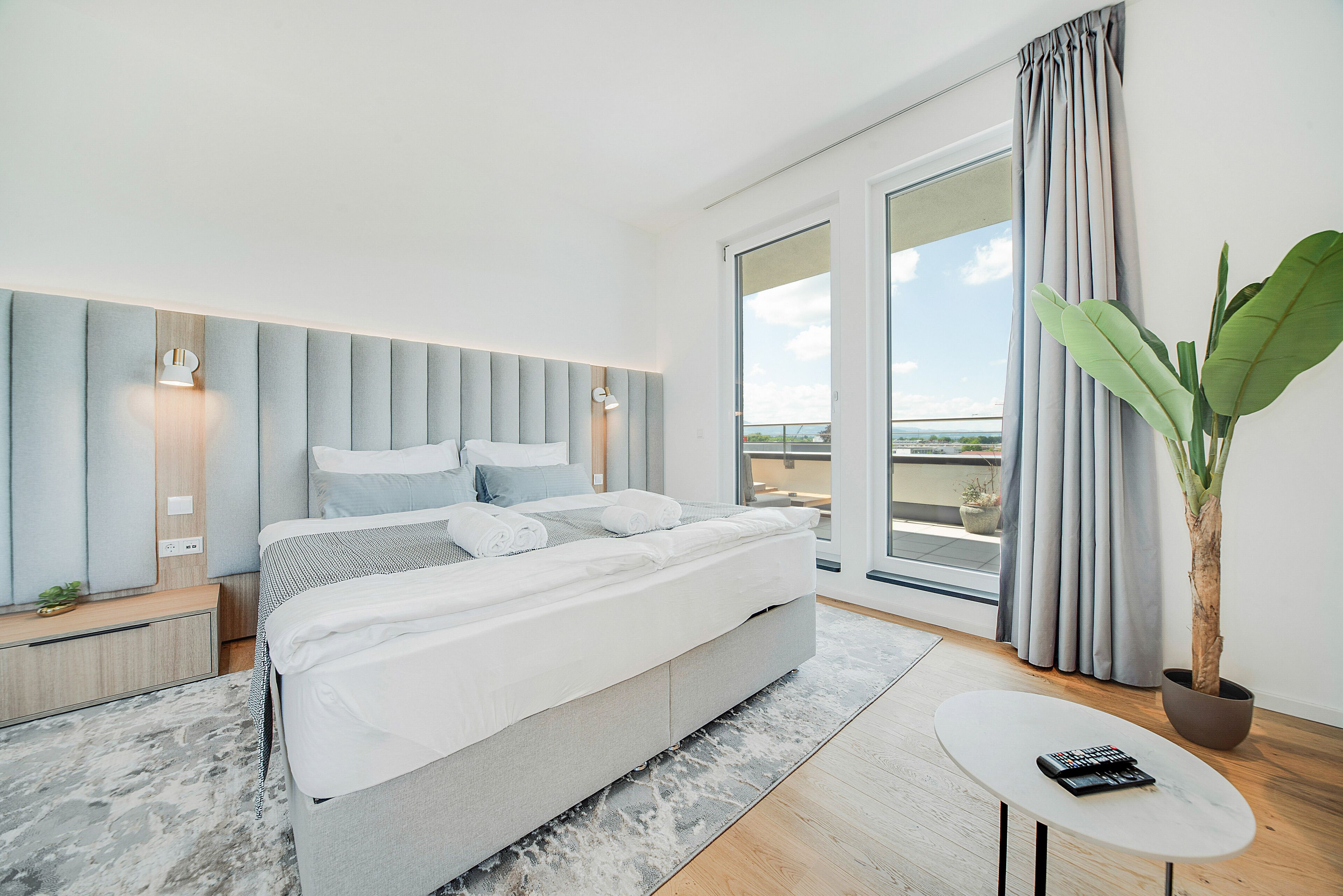 Un dormitorio moderno con una cama grande, una mesita, una planta en maceta y vistas al exterior.