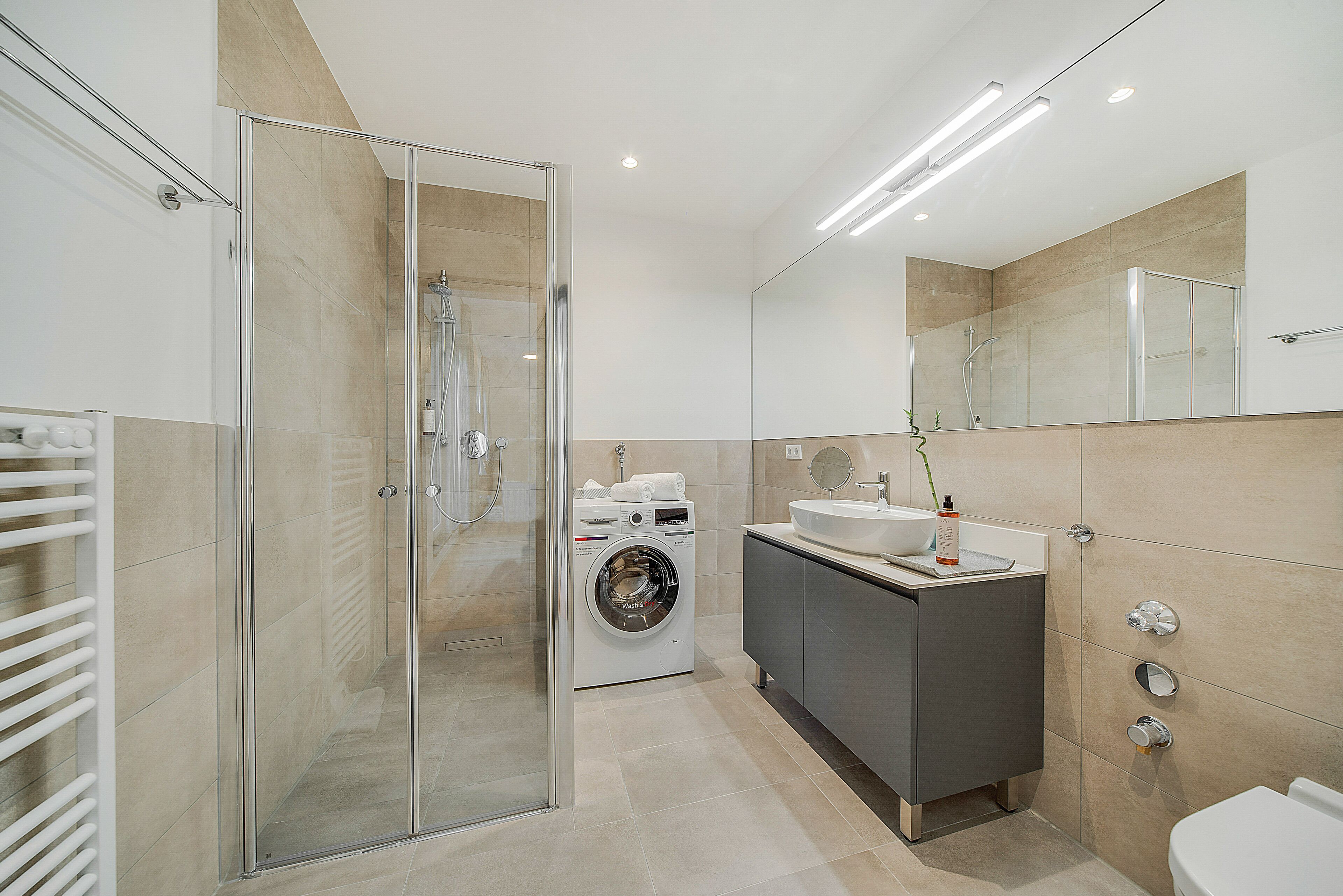 A modern bathroom with a glass shower enclosure, a built-in washing machine, a vanity with a sink, and a toilet.