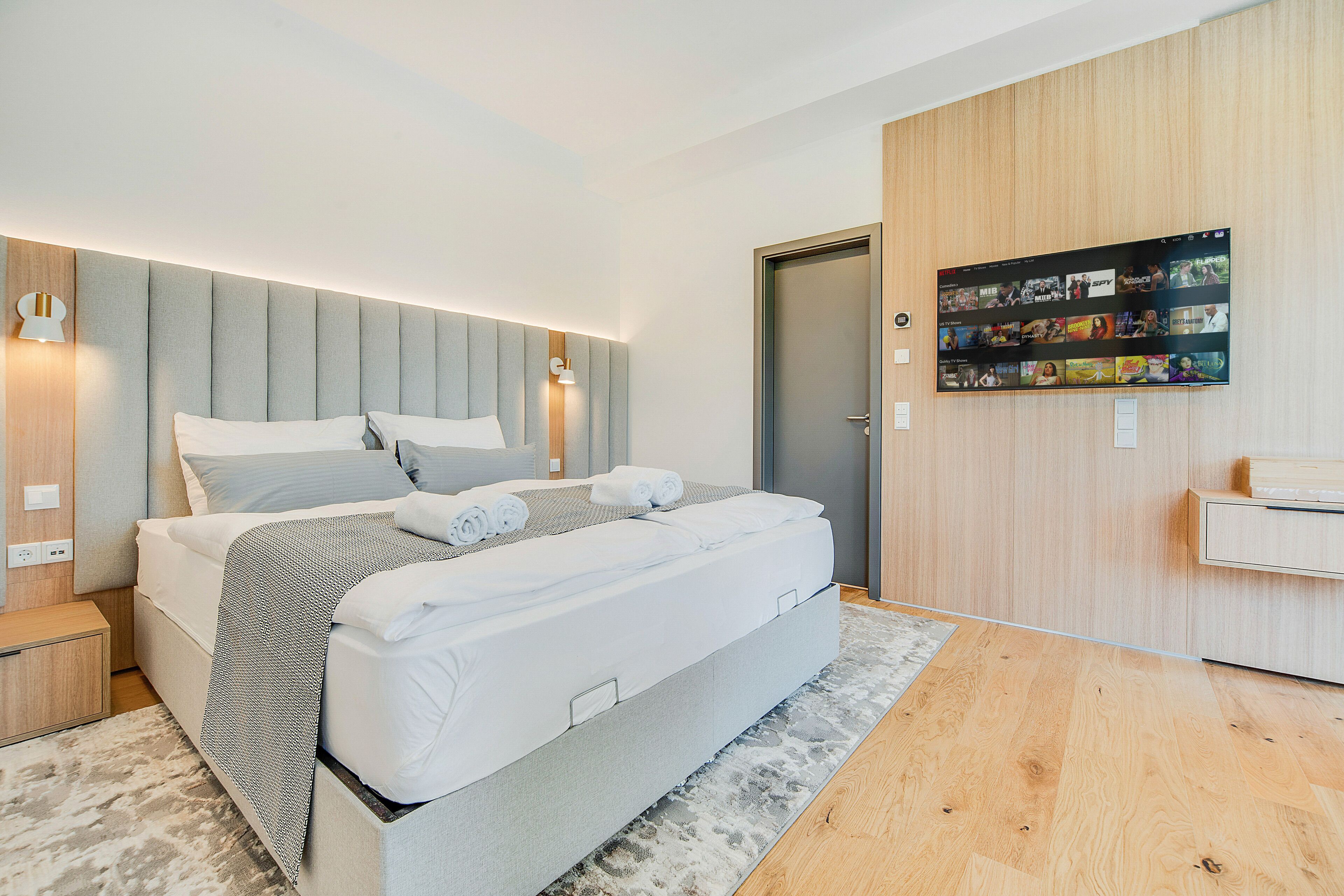 Un dormitorio moderno con una cama grande, un cabecero con iluminación integrada, un televisor de pantalla plana en la pared y piso de madera.