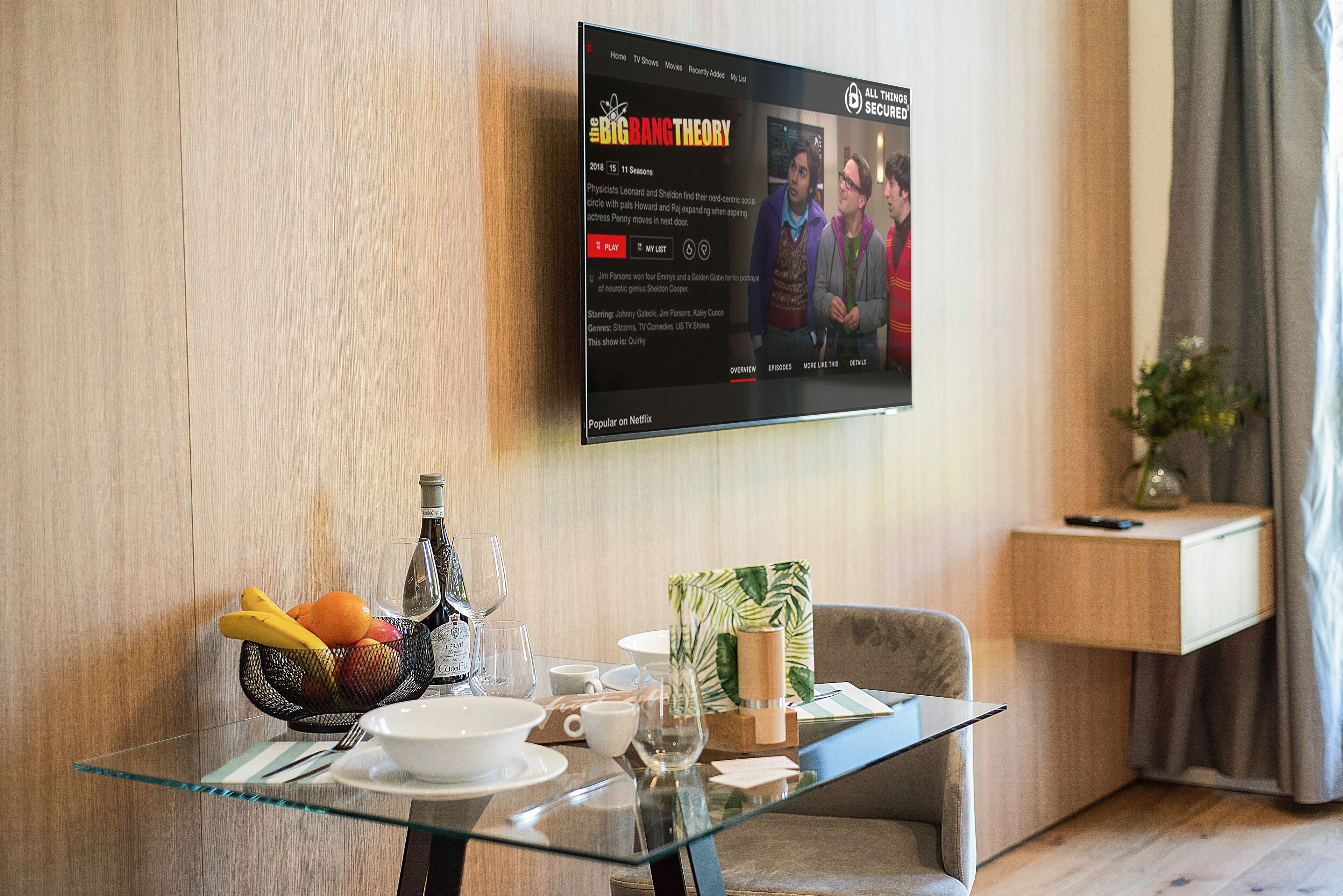 A hotel room with a glass table set for breakfast, a flat-screen TV mounted on the wall displaying a show, and a wooden nightstand with a vase.
