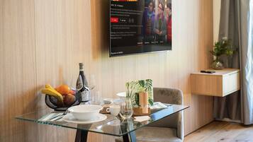 Ein Hotelzimmer mit Glastisch, gedeckt für das Frühstück, einem an der Wand befestigten Flachbildfernseher, der ein Programm zeigt, und einem Nachttisch mit einer Vase.
